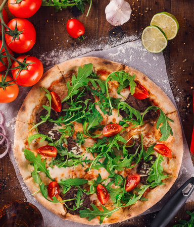Delicious pizza with arugula, tomatoes and meat freshly baked and served on wooden table surrounded by ingredientsの写真素材