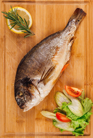 Grilled dorado fish lying on wooden cutting board with vegetables and lemon