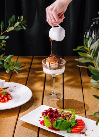 Person drizzling sauce on ice cream among a spread of assorted elegant dessertsの写真素材
