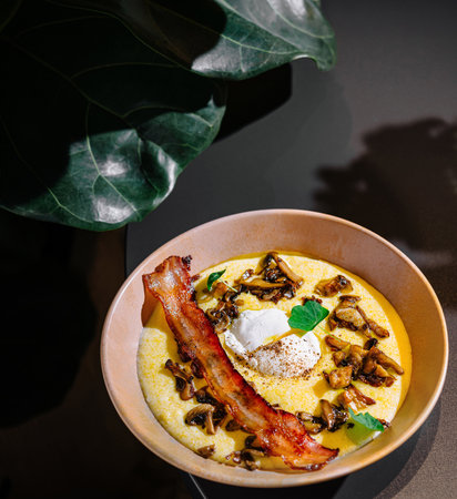 Delicious polenta with roasted mushrooms, crispy bacon and poached egg, seasoned with fresh herbs, served on a tableの写真素材