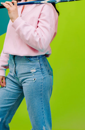 Woman holding a badminton racket on a green background is wearing jeans and a pink sweatshirt