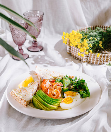 Plate with keto breakfast with shrimps, avocado, salad and eggs is served on a table with white...