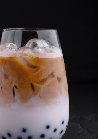 Glass filled with refreshing iced coffee mixed with milk and boba balls over black background