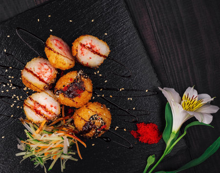Delicious fried sushi balls being served on a black stone plate decorated with sauce, sesame seeds and a flowerの写真素材
