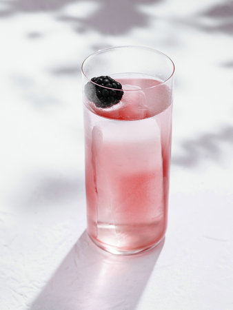 Tall glass with a refreshing pink cocktail, decorated with a blackberry and ice cubes, stands on a white background with contrasting shadowsの写真素材