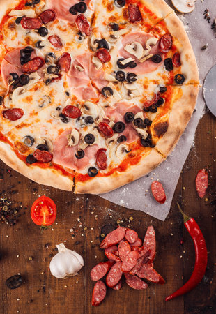Mouthwatering pizza with melted mozzarella, ham, mushrooms, and black olives, lying on a rustic wooden table surrounded by fresh ingredientsの写真素材