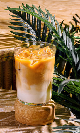 Refreshing iced latte beverage sitting on a wooden surface with green tropical plants and dappled sunlightの写真素材