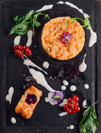 Salmon tartare being served on a black slate plate decorated with herbs, flower petals, and redcurrantの写真素材