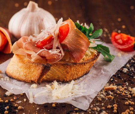 Toasted bread slice topped with prosciutto, parmesan, arugula and tomatoes laying on a wooden tableの写真素材