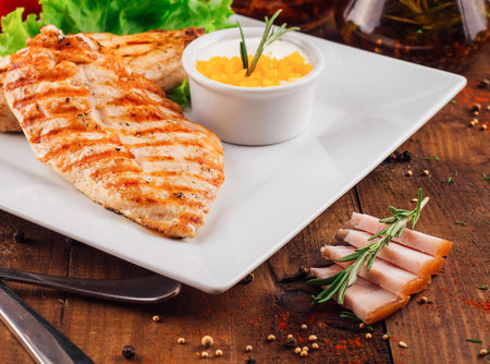 Two grilled chicken breasts served on a white plate with lettuce, sauce, and spicesの写真素材