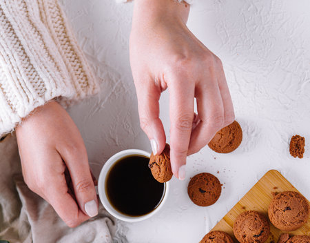 Close-up of hands holding a cookie with a cup of coffee and a plate of cookies on a white tableの写真素材
