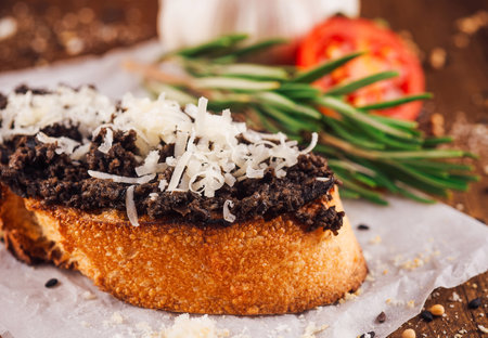 Delicious bruschetta is laying on white paper on a wooden table, with black truffle cream and parmesan cheese on topの写真素材
