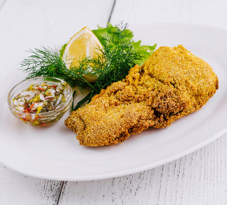 Breaded fish fillet served on a white plate with a slice of lemon, dill, lettuce, and a small bowl of sauce on the sideの写真素材