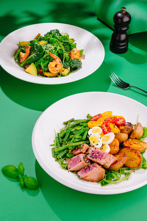 Two white plates with tasty fresh salads with shrimps, arugula, tomatoes and eggs on green backgroundの写真素材