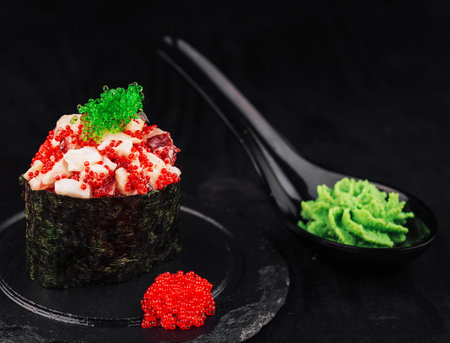 Gunkan maki sushi topped with tobiko and red caviar is presented on a slate plate, accompanied by wasabi and a pink flowerの写真素材