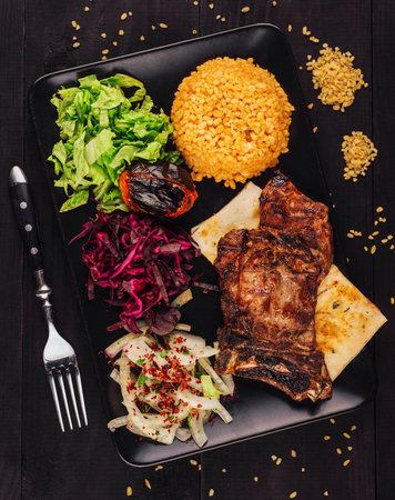 Grilled beef steak served on black plate with vegetables, shot from above on dark wooden background