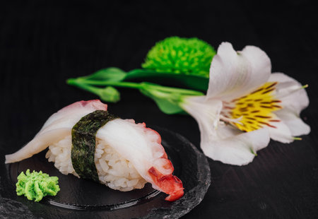 Single octopus sushi with a side of wasabi is laying on a black slate plate, with soy sauce and a flower in the backgroundの写真素材
