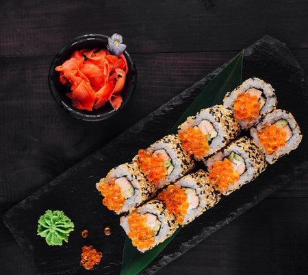 Sushi rolls set with red caviar and sesame seeds served on black stone slate on dark wooden table with ginger, wasabi and white orchidの写真素材