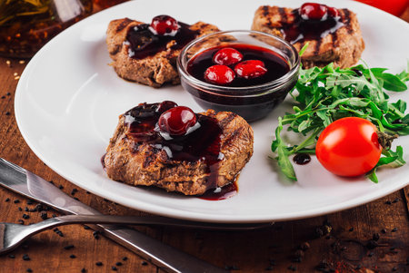 Grilled beef medallions with cherry sauce are served with arugula and cherry tomatoes on a white plateの写真素材
