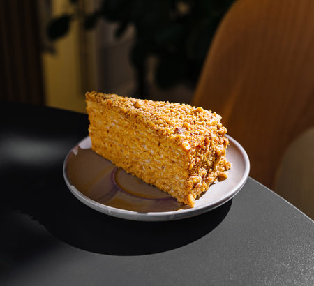 Slice of honey cake is sitting on a plate on a table in a cafe, illuminated by warm sunlightの写真素材