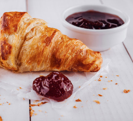 Delicious golden croissant is lying on white wooden surface with jam asideの写真素材