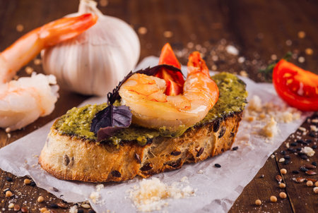 Toasted slice of bread with pesto sauce, grilled shrimps, tomato and basil leaf is laying on a wooden tableの写真素材