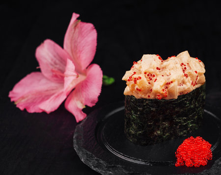 Fresh sushi with spicy sauce, decorated with red caviar, is served on a black stone plate next to a pink flowerの写真素材