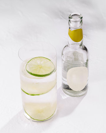 Tall glass filled with sparkling water and lime slices is standing on a white tablecloth next to a bottle with a blank labelの写真素材