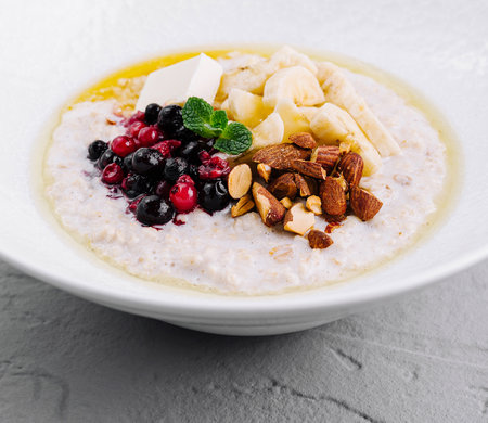 Oatmeal porridge is topped with berries, almonds, banana slices and mint in a white bowl for a healthy breakfastの写真素材