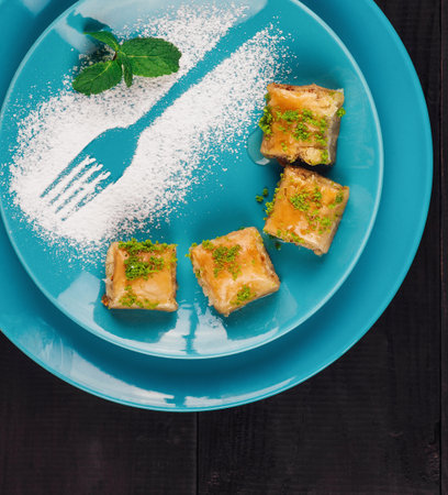 Delicious baklava with pistachios arranged on a blue plate with powdered sugar forming a fork, on a black wooden tableの写真素材