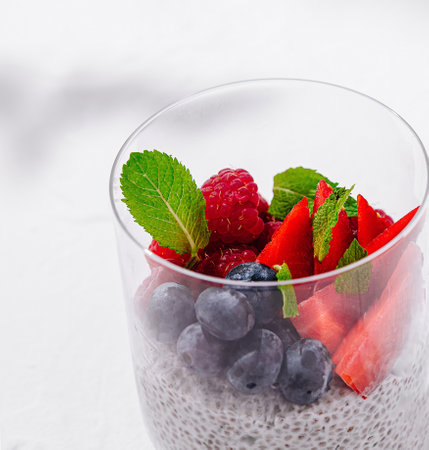 Delicious and healthy chia pudding with fresh raspberries, blueberries, strawberries and mint leaves in a glass on a white tableの写真素材