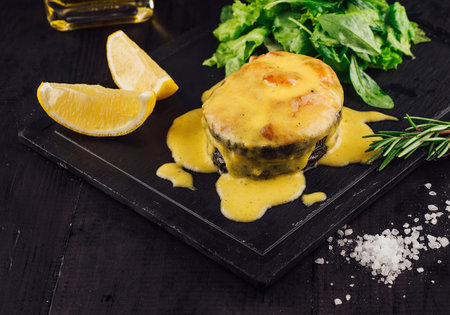 Delicious roasted fish with cheese sauce lying on black stone plate, decorated with lemon and herbsの写真素材