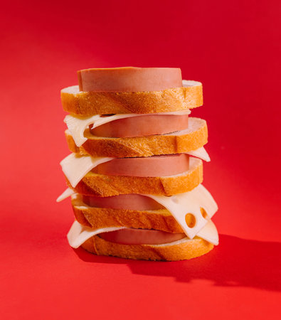 Precarious sandwich tower made of white bread, cheese, and bologna is standing on a red backgroundの写真素材