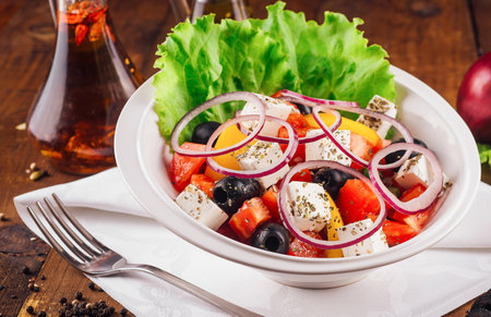 Traditional greek salad with fresh vegetables, feta cheese and olives on a white plate on a rustic wooden tableの写真素材