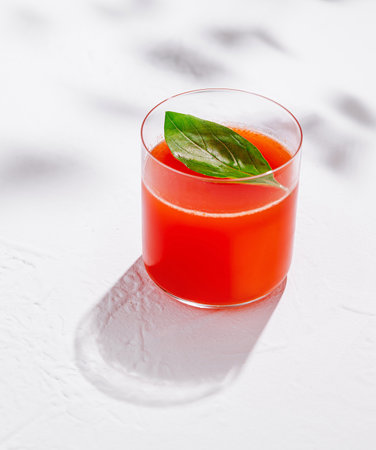 Glass of freshly squeezed tomato juice with basil leaf is standing on white table in harsh sunlight with shadowsの写真素材