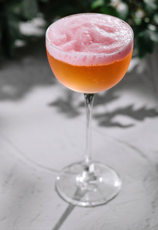 Stylish pink cocktail with a frothy top, served on a sleek, textured background with greeneryの写真素材