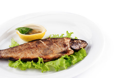 Grilled trout is lying on a bed of lettuce on a white plate, served with lemon and condiment