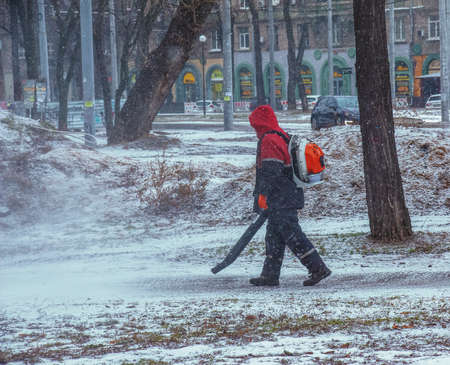 Seasonal work of city utilities in the park. A worker with a motorized backpack blower blows snow from a park path.の写真素材