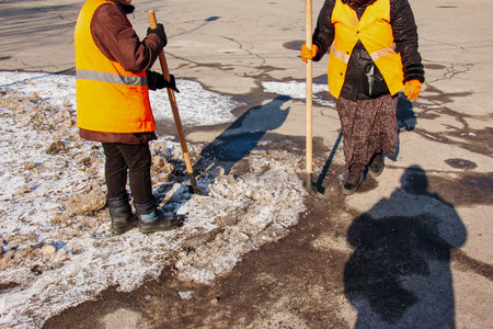 A woman worker cleans the ice and removes snow from paving slabs using an icebreaker. A man breaks ice with a steel blade crusher, an ice-breaking tool. The janitor cleans up the area.の写真素材