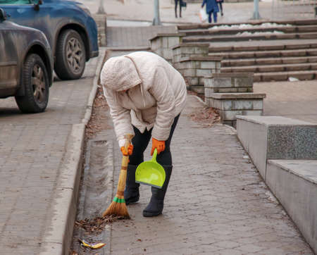 A worker with a broom sweeps the street from dust and debris. The janitor cleans the road. A female cleaner sweeps the sidewalk with a broom and dustpan.の写真素材