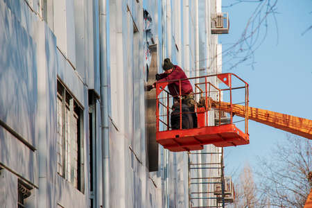 Dnepropetrovsk, Ukraine - 02.09.2022: Builder in a crane basket repairs the coating of a metal structure on the wall of the facade of a building.のeditorial素材