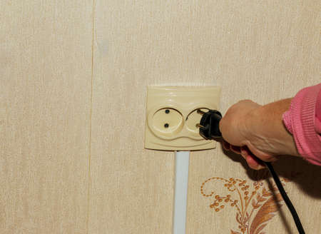 Close-up of a woman's hand holding a black plug and about to plug it into a white socket on the wall of a house. energy saving conceptの写真素材