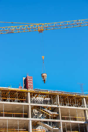 A crane lifts concrete for a modern metal-concrete structure of a residential building. Container with concrete against the blue sky.の写真素材
