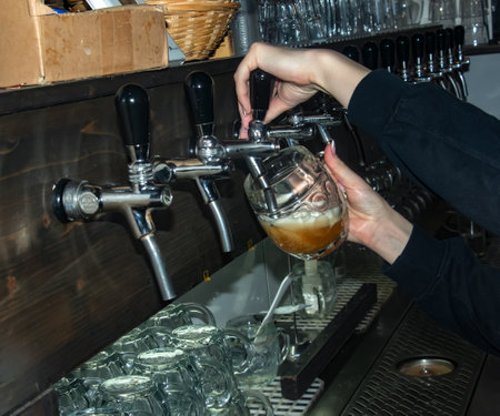 The hand of a bartender girl at a beer tap pours draft beer into a glass served in a restaurant or...