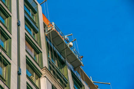 Suspended platform, construction cradle hanging on building. Installation and repair work. Building facades construction works. Facade of high-rise building under construction, finishing worksの写真素材