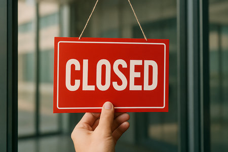 Hand holds a vibrant red 'CLOSED' sign against a blurred glass door, signaling business closure.の素材
