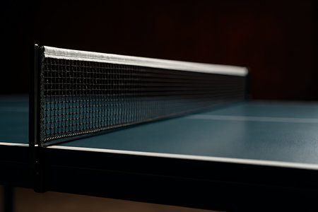 A dimly lit table tennis setup shows the net dividing the playing surface in a dark arena.の素材