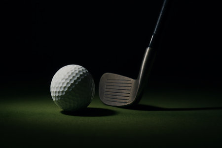 A white dimpled golf ball sits poised next to an iron club on green turf with a dark background.の素材
