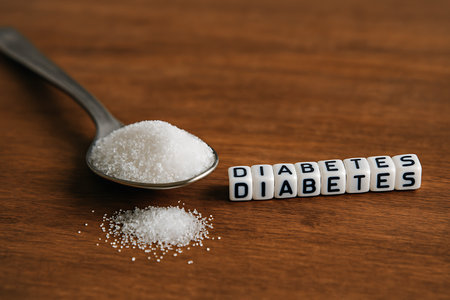A spoonful of granulated sugar beside letter dice spelling out diabetes on a wooden background.の素材