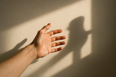 A persons hand reaches out into warm sunlight, creating a dynamic interplay of light and sharp geometric shadows on a textured wall.の素材